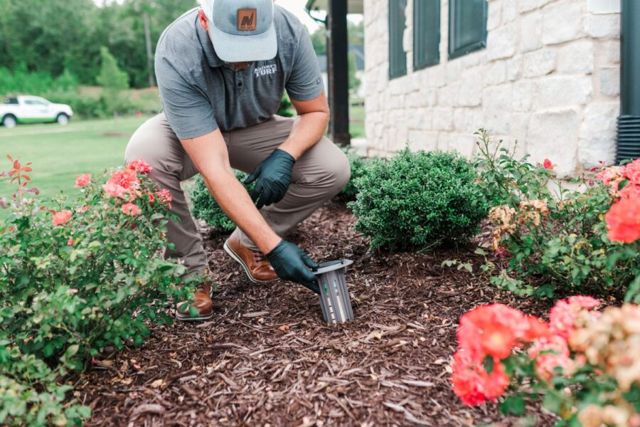 Nature's Turf employee inspecting a termite bait station in the natural area beside a white brick home