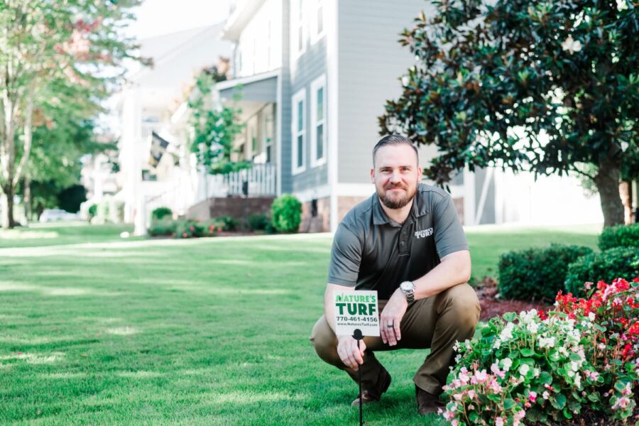 Nature's Turf employee squatting in lush green yard with a Nature's Turf sign beside some flowers