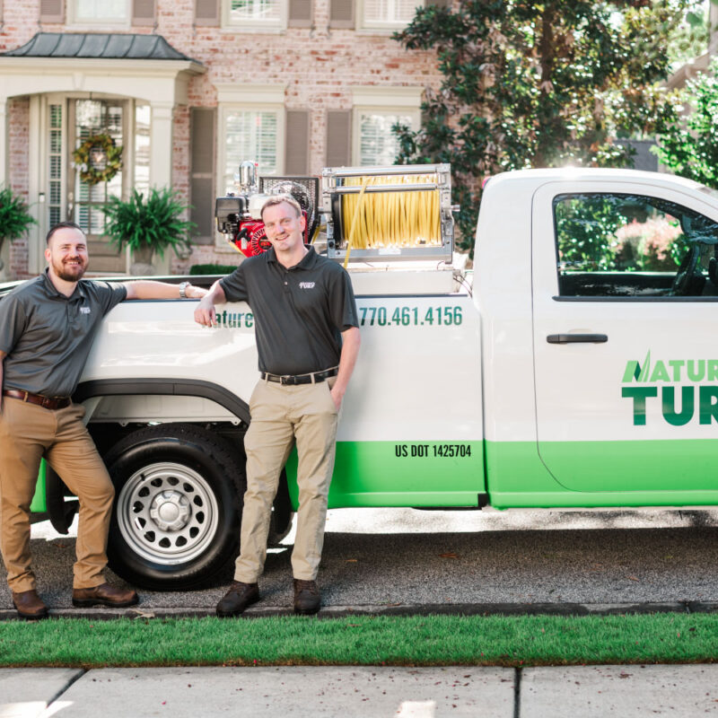 Nature's Turf employees standing by Nature's Turf truck in front of a brick house with a lush, green lawn