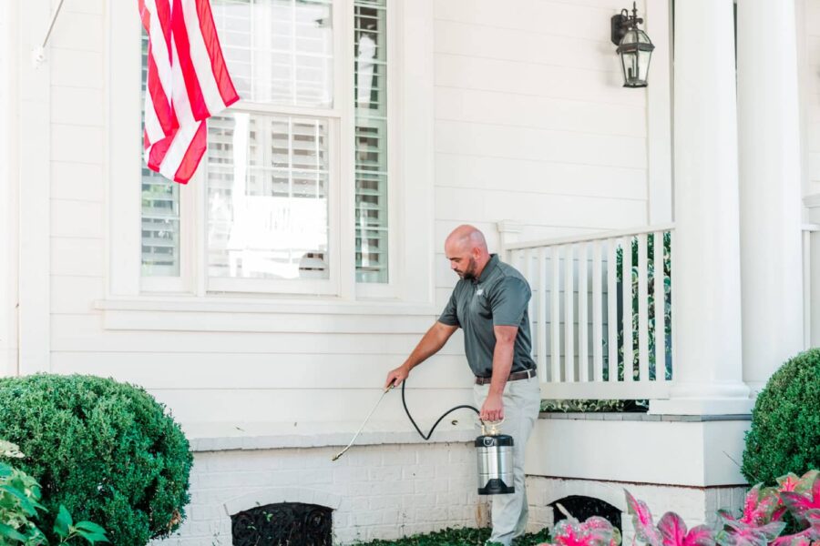 Nature's Turf employee spraying for cockroaches at the base of a beautiful white brick home with an American flag and surrounded by lush grass, shrubs, and flowers