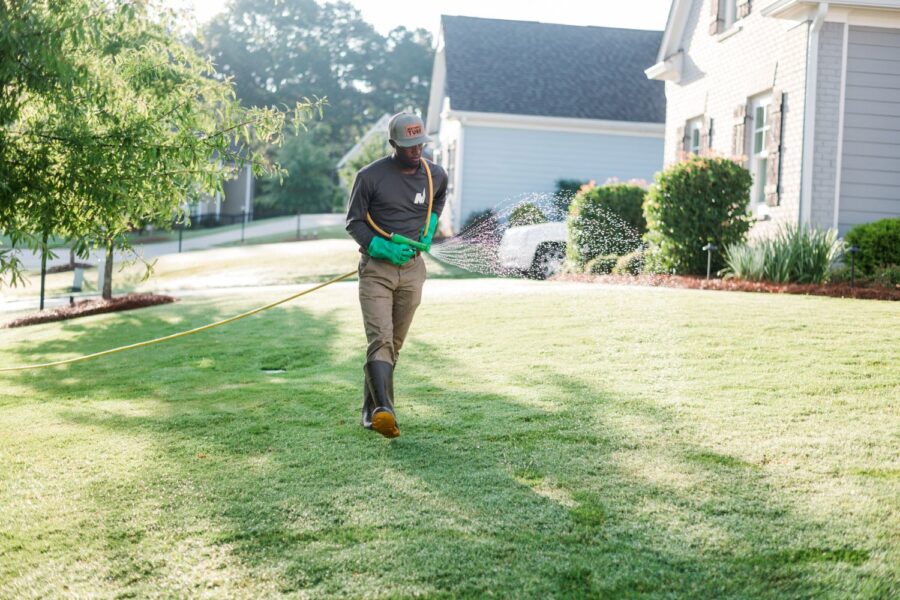 Nature's Turf employee walking on a green lawn in front of a house and carrying a hose that is spraying liquid lawn products