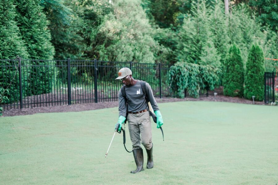 Nature's Turf employee spraying weed control products on a manicured green lawn surrounded by a black gate and mature trees.