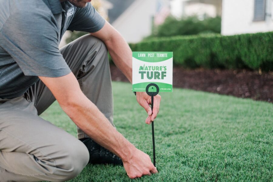 Nature's Turf employee planting company sign in a lush green lawn with hedges and a white home in the background