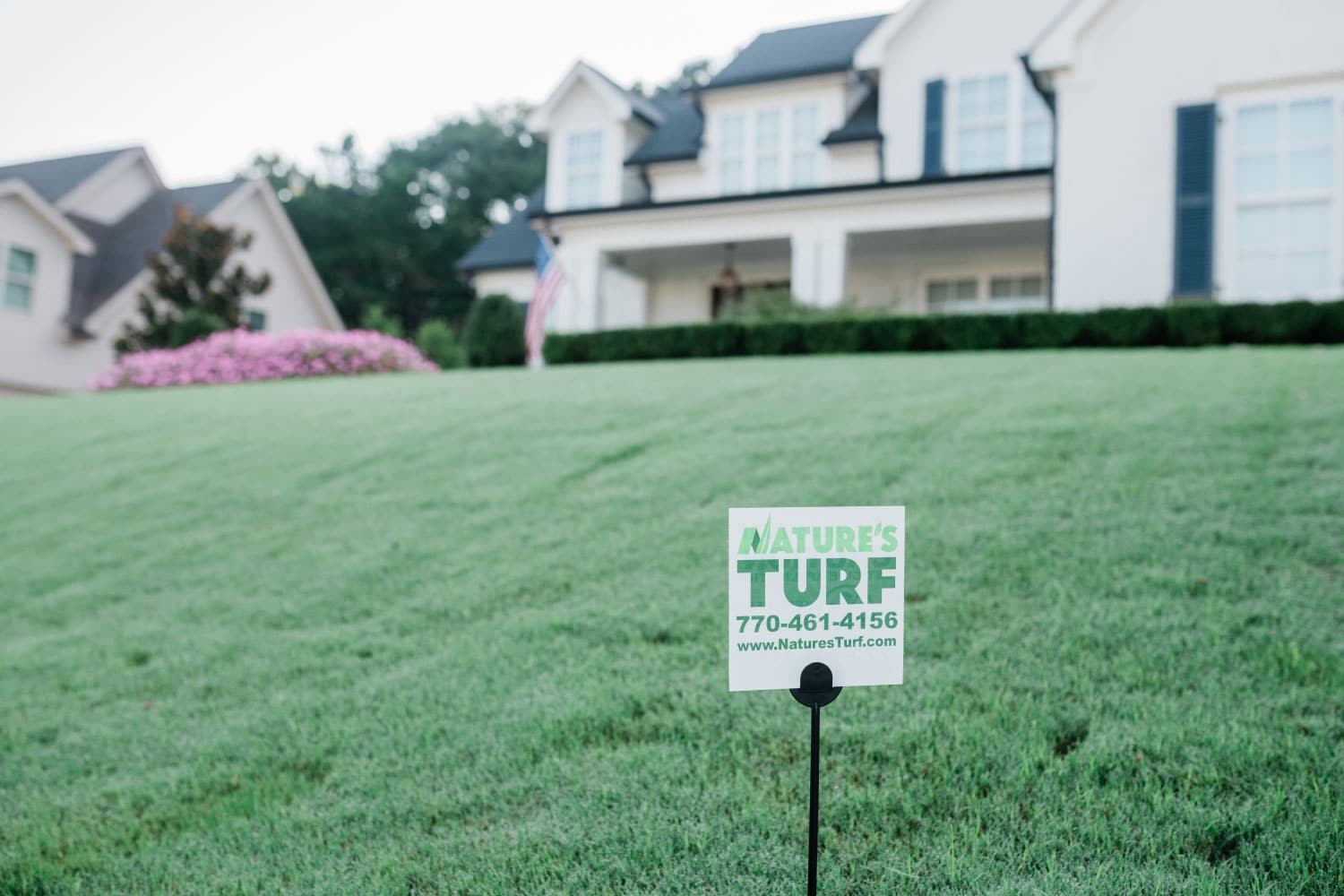 Nature's Turf yard sign in a lush, green lawn in front of a white, modern home with an American flag in front