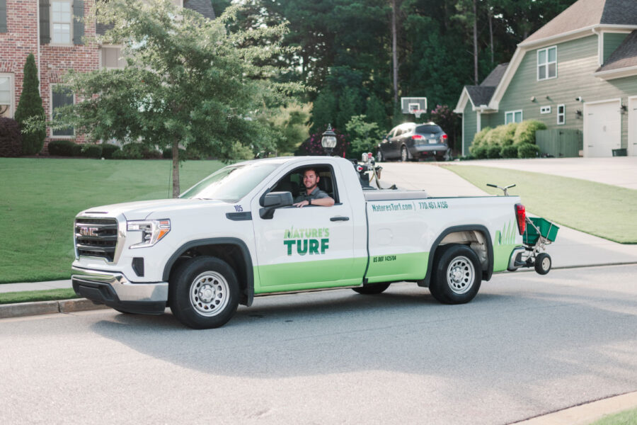 Nature's Turf employee sitting in a company truck and smiling in front of a brick home with a beautiful lawn
