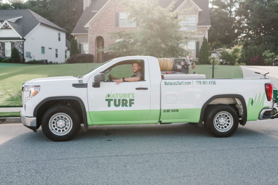 Nature's Turf employee smiling and sitting in the driver's seat of a Nature's Turf truck on the street in front of a brick home with a beautiful lawn