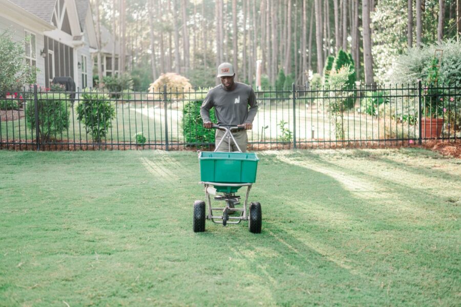 Nature's Turf employee spreading seed on a beautiful, green lawn in the backyard of a home with a wrought iron fence