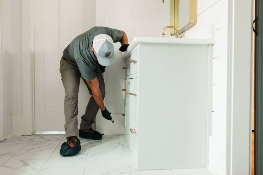 Nature's Turf employee spraying pest control products in a white residentail bathroom with a white marble floor, white cabinets, and gold fixtures
