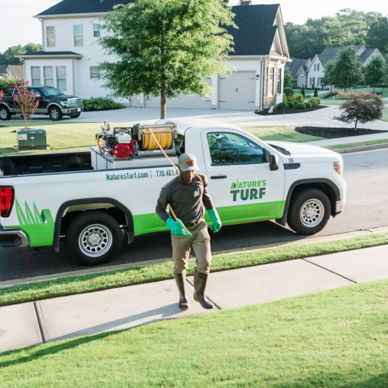 Nature's Turf employee in a uniform walking away from a company truck parked on a suburban street lined with beautiful single-family homes with manicured lawns
