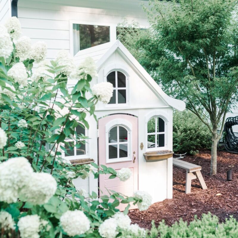 A children's playhouse in a mulched area next to a white house and surrounded by shrubs and white hydrangeas