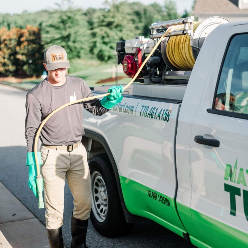 Nature's Turf employee standing between a street curb and the Nature's Turf truck and holding a weed control product applicator hose