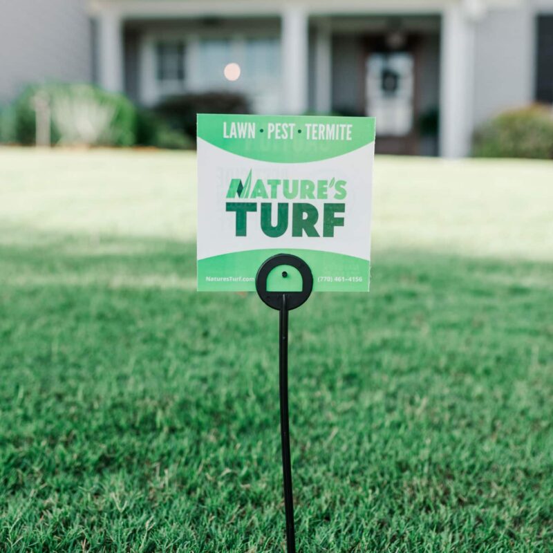Nature's Turf yard sign stuck in the middle of a lush green lawn with a big white house in the background