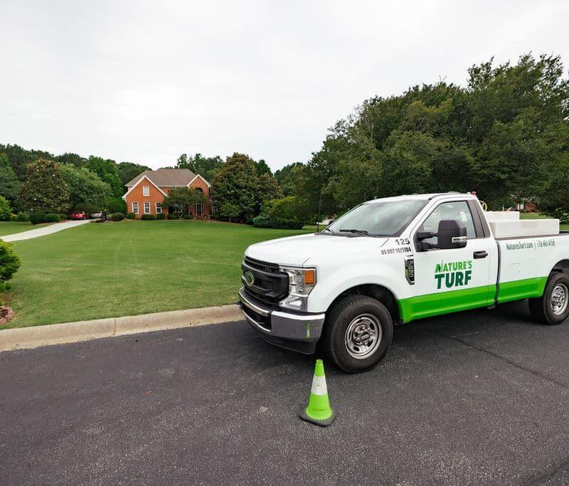 Nature's Turf company truck parked on the street in front of a brick home with a large beautiful lawn and a long driveway