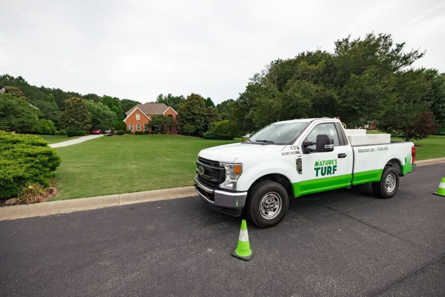 Nature's Turf company truck parked on the street in front of a brick home with a large beautiful lawn and a long driveway