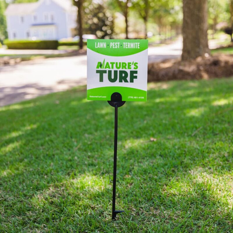 Nature's Turf yard sign stuck in the middle of a lush green lawn with a blurred-out street and a big white house in the background