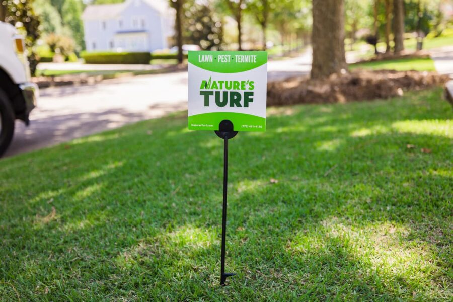 Nature's Turf yard sign stuck in the middle of a lush green lawn with a blurred-out street and a big white house in the background
